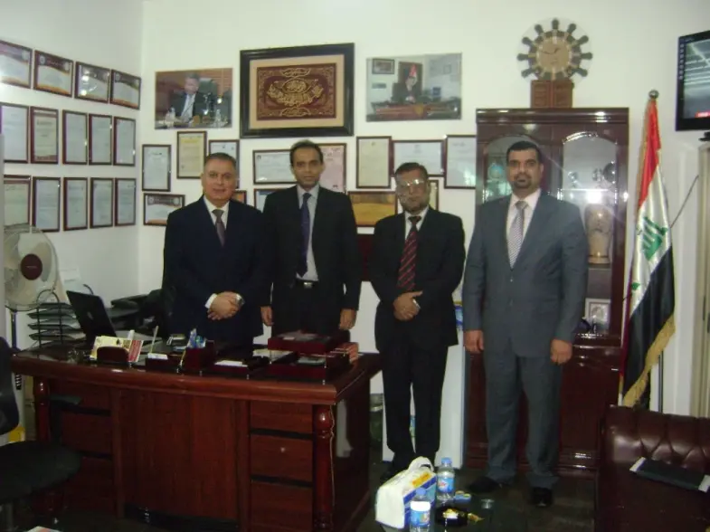 Four men standing in an office beside the Iraqi flag